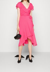 Robe portefeuille rose vif avec manches volantées et ourlet asymétrique. Sac à main noir avec chaîne dorée et escarpins pointus avec nœuds décoratifs.