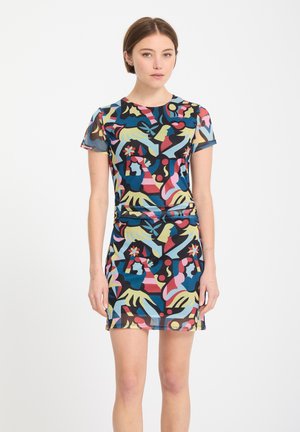 Jeune femme portant une robe à manches courtes avec un motif abstrait multicolore, debout devant un fond blanc uni.