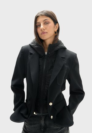 Joven mujer con cabello liso castaño, lleva pendientes dorados, blazer negro en capas y chaqueta acolchada, manos en los bolsillos, fondo neutro.