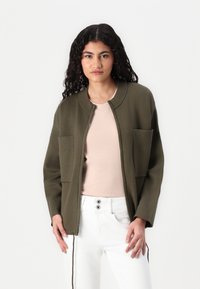 Olijfgroene jas met een ronde kraag, ritssluiting aan de voorkant, geribbelde accenten op de zakken en een zachte textuur. Draagt over een lichtroze shirt en witte broek.