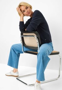 Marine ribbeltrui, lichtblauwe wijde jeans en witte sneakers. Zittend op een stoel met chromen frame, een gevlochten zitting en een zwarte rugleuning.