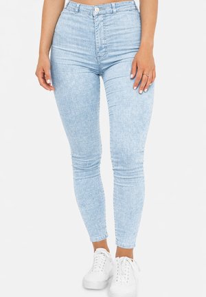 Kobieta ubrana w jasnoniebieskie, dopasowane jeansy z wysokim stanem i białe trampki, stojąca na tle jednolitej białej ściany.