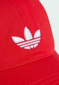 Sile hüübinud punane müts, mis on valmistatud siledast kangast, millel on valge brodeeritud kolme lehe logo ja kaks horisontaalset triipu. Kõver äär, kus on nähtavad õmblused.