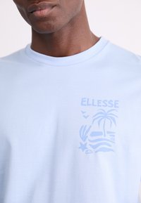 Šviesiai mėlynas medvilninis marškinėliai su toniniu grafikos dizainu, kuriame pavaizduoti palmės ir jūros kriauklės, o prekės ženklas "ELLESSE" užrašytas šviesiai mėlynu šriftu.