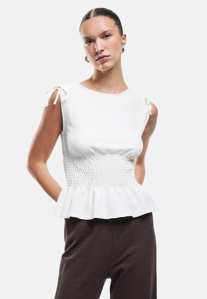 Witte mouwloze top met gestructureerde stof, gerimpelde taille en peplumrand. Heeft geknoopte accenten bij de schouders. Gepaarde met donkere broeken.