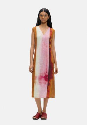 Kobieta z długimi ciemnymi włosami ubrana w sukienkę midi bez rękawów z dekoltem w serek i pionowymi panelami tie-dye w kolorze pomarańczowym, różowym, białym i czerwonym oraz czerwone klapki z odkrytymi palcami.