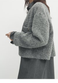 Cardigan en laine texturée grise avec une coupe ample, doté d'une poche poitrine et d'un détail de bouton, superposé sur une chemise grise foncée.