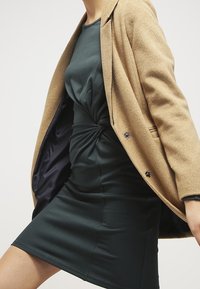 Robe verte avec un détail de nœud tordu à la taille, portée sous un manteau en laine beige avec des accents de boutons et une doublure intérieure lisse.