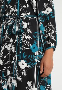 Blommig klänning i svart med vita och teal-mönster, med knytmidja, knappdetaljer och halvärmar med elastiska manschetter.