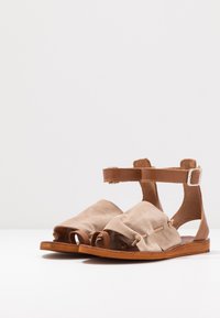 Beige suede sandals with an ankle strap and brown leather accents. Features open toes, gathered fabric detail, and flat wooden soles.