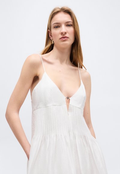 Blondínka v bielych šatách bez rukávov s tenkými ramienkami a malým zapínaním na hlbokom výstrihu do V, stojaca pred jednoduchým pozadím.