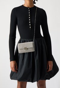 Svart ribbad långärmad klänning med en henley-krage och metallknappar; kombinerad med en silverfärgad crossbody-väska prydd med strass.