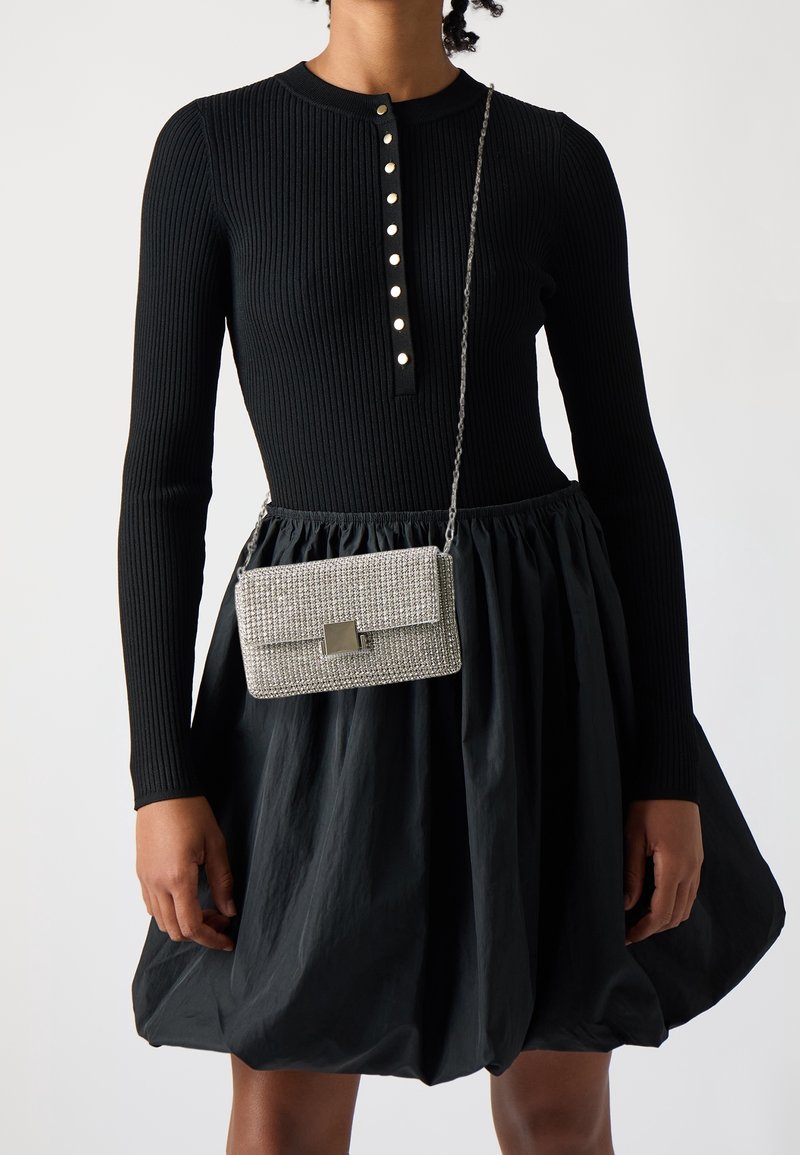 Svart ribbad långärmad klänning med en henley-krage och metallknappar; kombinerad med en silverfärgad crossbody-väska prydd med strass.