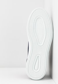 Semelle d’une basket blanche avec un motif de bande de roulement ondulé, positionnée verticalement dans un angle blanc entre un mur et le sol.