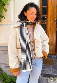 Veste en polaire crème avec des accents marron, fermeture à boutons et deux poches avant. Écharpe rayée noire et blanche, jeans bleu clair, top en dentelle en dessous.