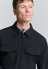 Zwarte jas met een ritskraag, twee borstzakken en een zachte textuur. Beschikt over een lichtpaarse binnenkraag en een strak ontwerp.