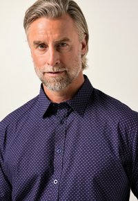 Camicia button-up viola scuro con un motivo geometrico in tonalità più chiare. Il tessuto appare liscio con un colletto strutturato e una leggera lucentezza.