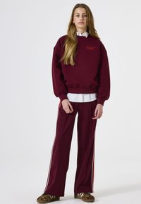 Sudadera color burdeos con mangas abullonadas, camisa blanca debajo y pantalones burdeos a juego con franjas laterales, combinados con calzado estampado.