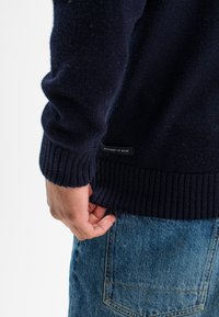 Navy gebreide trui met geribde manchetten en zoom; label "BUTCHER OF BLUE" zichtbaar aan de linkerkant in de buurt van de onderkant. G gedragen met denim jeans.