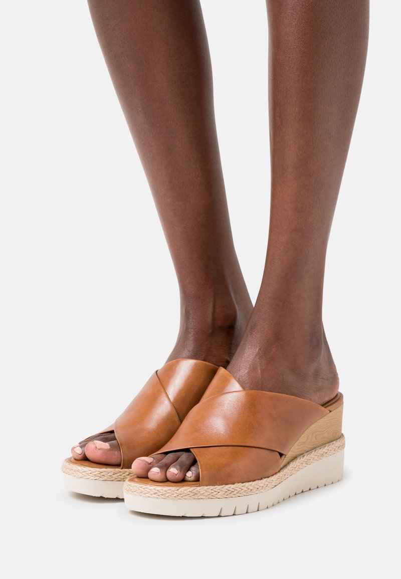 Sandales compensées en cuir marron avec des brides croisées portées sur des pieds à peau foncée sur fond blanc.