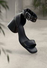 Schwarze High-Heel-Sandale mit quadratischer Zehenpartie, zwei breiten Riemen und einem Knöchelriemen, der mit metallischen Nieten und einem Schnallendetail versehen ist.