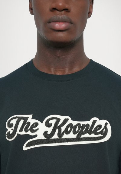 Melns kokvilnas T-krekls ar izteiksmīgu baltu un melnu izšūtu zīmola logotipu "The Kooples" uz krūtīm. Klasisks apaļās kakla dizains.