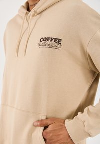 Haut à capuche beige avec le texte "HARMONIE DU CAFÉ" sur la poitrine, cordons de serrage, poche kangourou, et main reposant à l'intérieur de la poche.