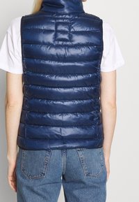 Marineblaues Steppweste mit hohem Kragen, gestepptem Design und glänzender Textur; über einem weißen Hemd getragen, kombiniert mit Jeans.