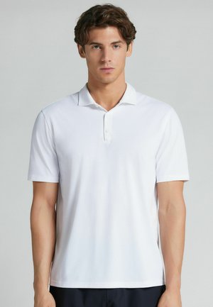 Joven de pie, vistiendo una camiseta polo blanca lisa de manga corta, mirando hacia adelante sobre un fondo gris claro.