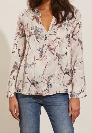 Femme portant un chemisier crème à imprimé floral et oiseaux grue, associé à un jean bleu, debout devant un fond neutre.