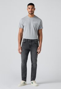 Mees seisab näoga ettepoole, kandes halli Levi's T-särki, musti teksaseid ja valgeid tossusid roheliste triipudega, taustaks lihtne hele pind.