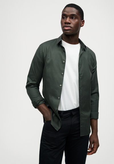 Άνδρας με σκούρο πράσινο πουκάμισο, φορεμένο πάνω από λευκό μπλουζάκι, και μαύρο παντελόνι, στέκεται με το ένα χέρι στην τσέπη, κοιτάζοντας πλάγια.