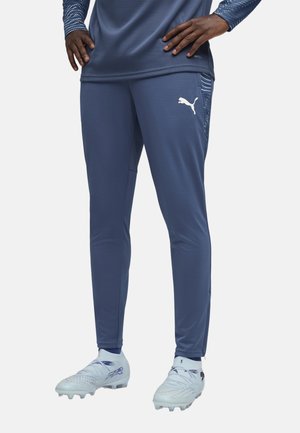 Osoba ubrana w niebieskie sportowe spodnie Puma i dopasowaną bluzę z długim rękawem, stojąca z rękami na biodrach, ubrana w jasnoniebieskie korki.