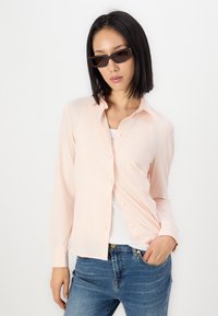 Soe roosa nööpidega särk, millel on krae ja pikad varrukad, valmistatud siledast kangast, koos siniste teksapükste ja tumedate prillidega.