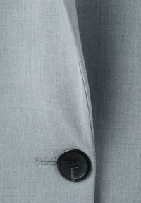 Nærbillede af en ærme på en grå blazer, der viser en sort firhulsknap fastgjort gennem en syet knaphul.