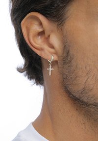 Silvrig hoopörhänge med en liten, texturerad korsanhängning som hänger från det, bärs i vänster öra. Hudton synlig runt örhänget.