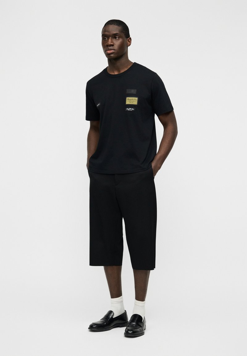 T-shirt en coton noir avec des patchs rectangulaires, associé à un short ample noir. Le mannequin porte des mocassins noirs et des chaussettes blanches.