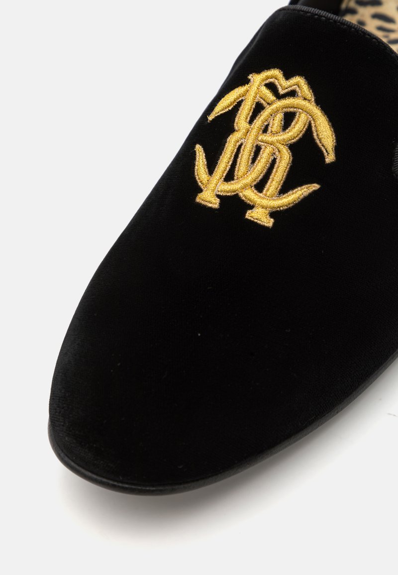 Zapato negro de terciopelo tipo slip-on con un logotipo bordado en oro elevado. El interior presenta un patrón de estampado de leopardo.