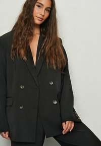 Mustat kaksiriviset bleiserit, joissa on avonainen kaulus, metallipainikkeet ja rento istuvuus. Teksturoitu kangas, jossa on sileä pinta.