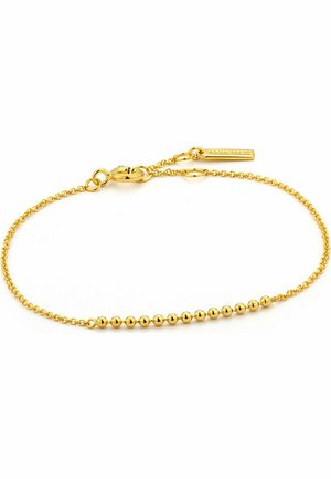 Goldarmband mit kleinen runden Perlen entlang einer feinen Kette, ausgestattet mit einem Karabinerverschluss und einem Markenanhänger. Glatte, glänzende Oberfläche.