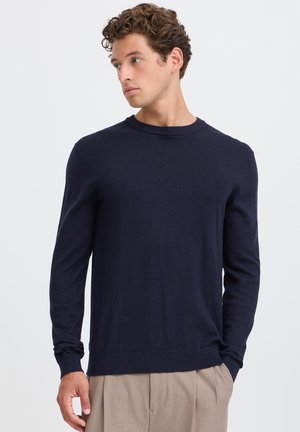 Jonge man met krullend haar die een marineblauwe lange mouwen trui en beige broek draagt, kijkt naar links tegen een simpele achtergrond.
