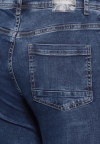 Dark blue denim jeans with a back pocket, faded texture, and contrasting stitching. Features a tag at the waistband with a floral design.