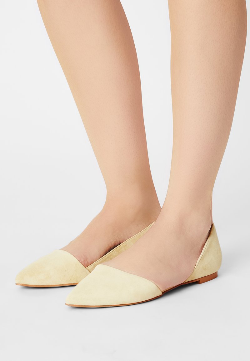Pieds portant des chaussures plates beige clair à bout pointu avec un petit talon sur un fond gris clair.