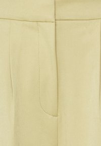 Calças beje ajustadas com uma textura suave, pregas frontais e bolsos laterais com detalhes de costura discretos.