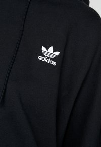 Svart tröja med huva, med en vit broderad Adidas-logotyp och tre vita ränder. Mjuk textur, synliga sömmar.