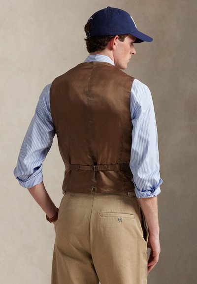 Καφέ σατέν γιλέκο πάνω από ένα ανοιχτό μπλε και λευκό ριγέ πουκάμισο, συνδυασμένο με χακί παντελόνια. Διαθέτει πόρπη στην πλάτη και μανίκια που έχουν γυριστεί προς τα πάνω.