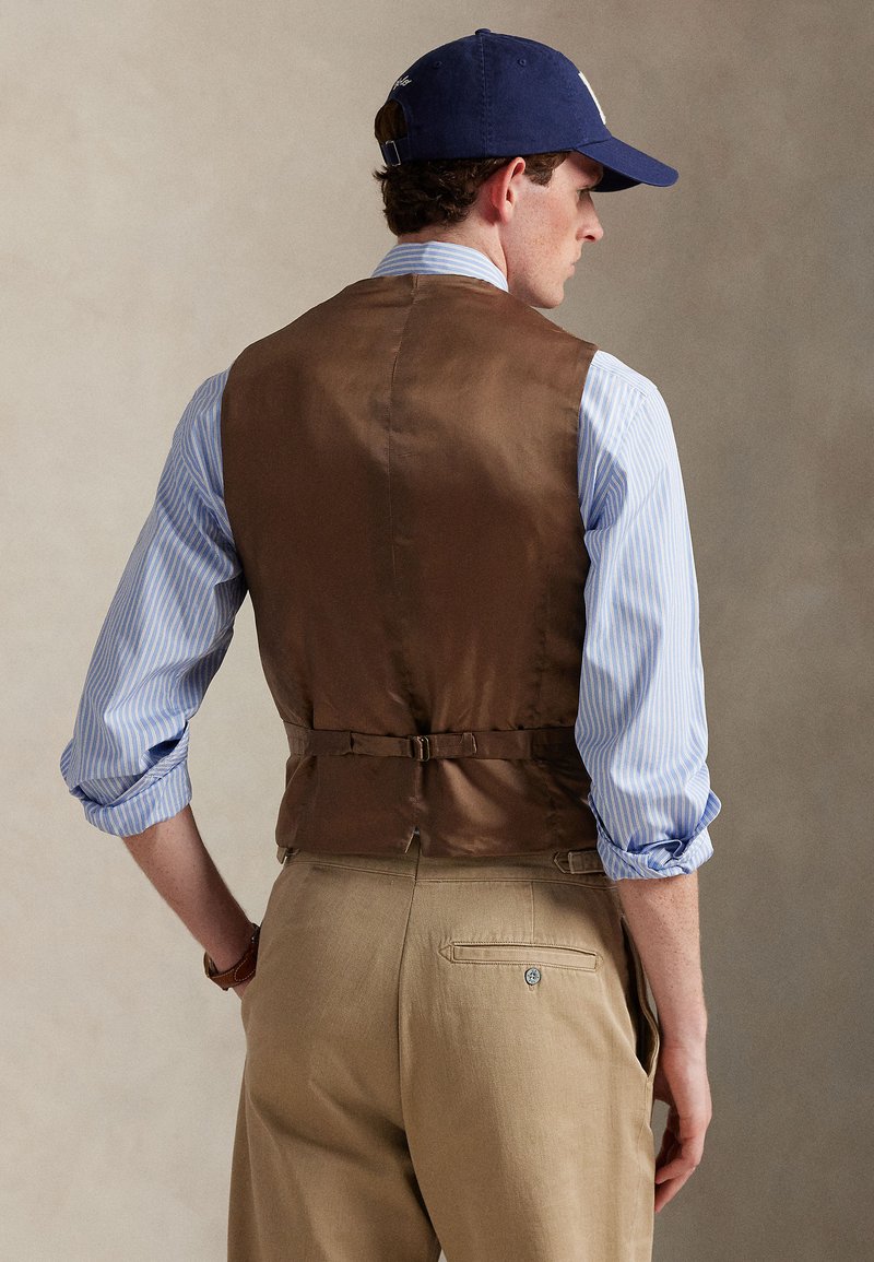 Gilet en satin marron porté sur une chemise rayée bleu clair et blanc, associé à un pantalon kaki. Caractéristiques : boucle au dos et manches retroussées.