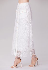 Jupe maxi en dentelle blanche avec un devant boutonné, deux poches à rabat et un ourlet festonné. Associée à des sandales à talons hauts blanches à lanières.