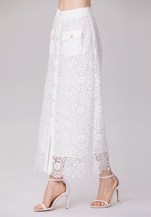 Jupe maxi en dentelle blanche avec un devant boutonné, deux poches à rabat et un ourlet festonné. Associée à des sandales à talons hauts blanches à lanières.