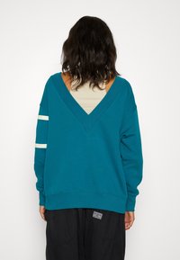 Tealfärgad sweatshirt med vid V-ringning och trekvartsärmar. Har vita horisontella ränder på ena ärmen och en avslappnad passform.